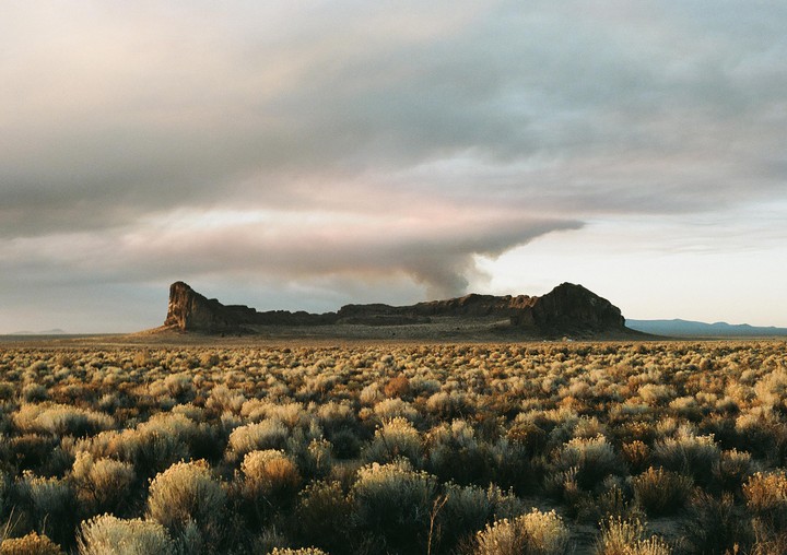 A brush fire burns behind Fort Rock, a large caldera in Bend, Oregon, with a field of desert brush in the foreground.