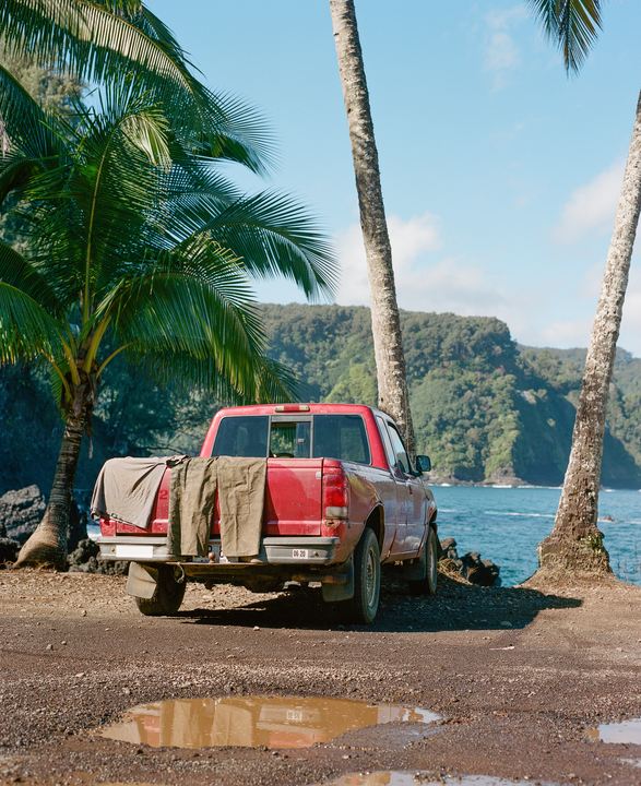 A red pickup truck is parked on a dirt road next to the ocean, with palm trees and lush green mountains in the background.