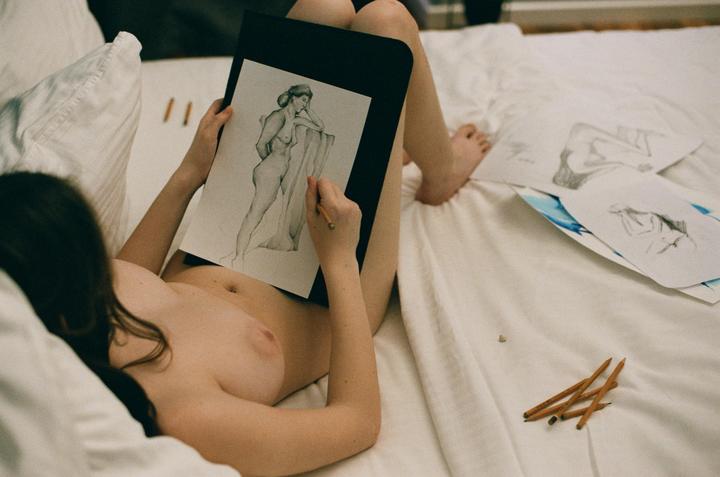 A woman lies on a bed drawing a nude figure with a pencil.