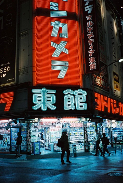 A person with an umbrella walks past a brightly lit store with neon signs at night.