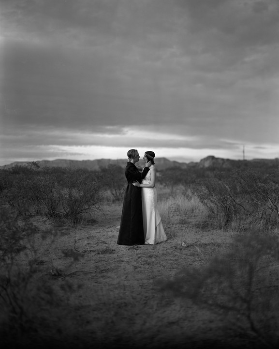Two women embrace in a desert landscape under a cloudy sky.