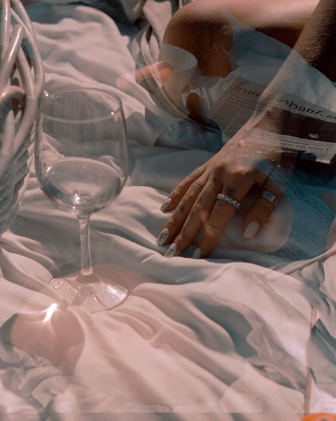 A double exposure shows a hand with rings and painted nails next to a wine glass on a white sheet, with a newspaper and a person's leg visible in the background.