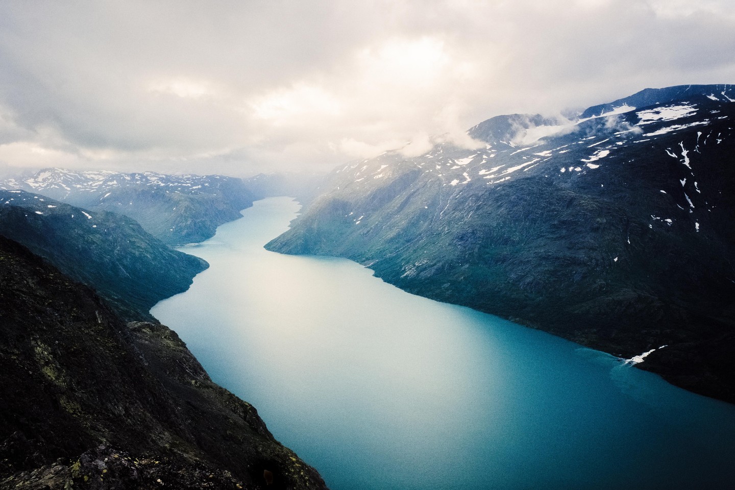 A long, narrow lake winds through a valley surrounded by mountains with patches of snow under a cloudy sky.