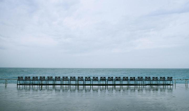 A row of empty chairs sits on a wet surface facing the ocean under a cloudy sky.