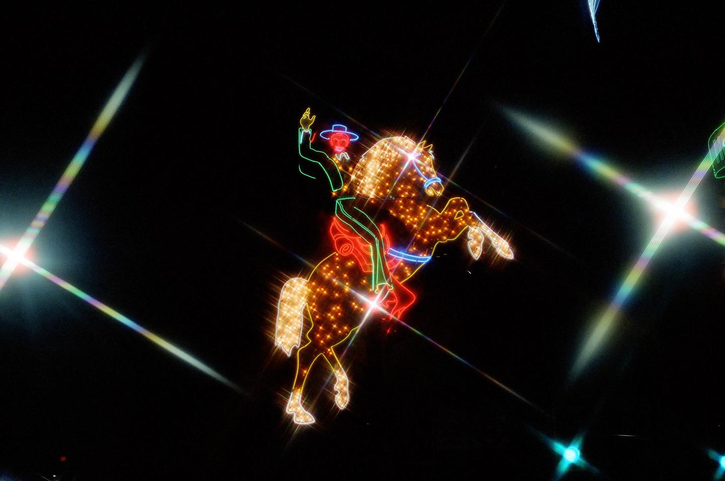A neon cowboy on a rearing horse is illuminated against a dark background with starburst light flares.