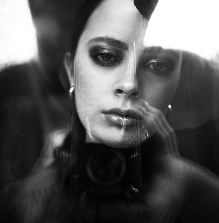A woman with dark hair and makeup looks directly at the viewer, with a camera visible in a double exposure.