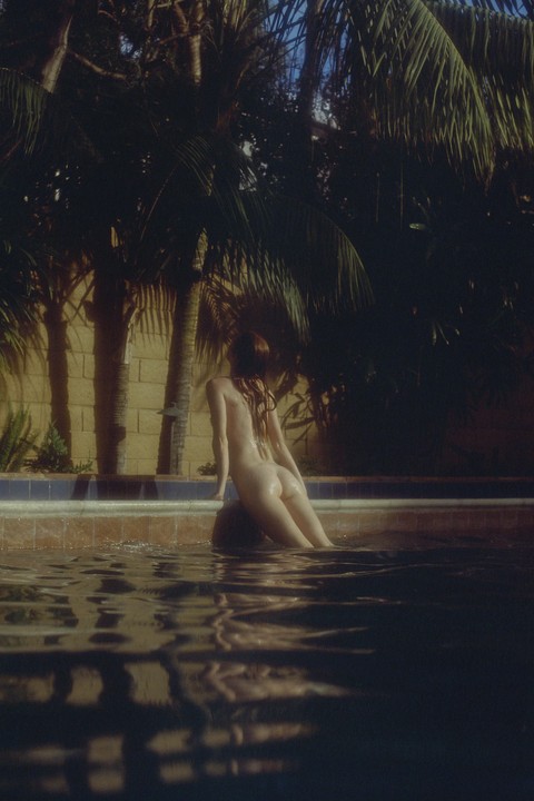 A nude woman sits on the edge of a swimming pool with palm trees in the background.