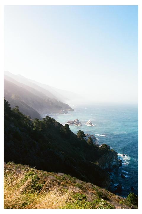A hazy view of the California coast shows steep, forested cliffs meeting the ocean with rocky outcrops.