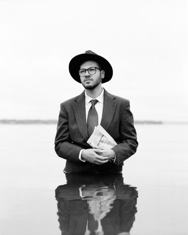 A man in a suit, hat, and glasses stands in water holding a newspaper.