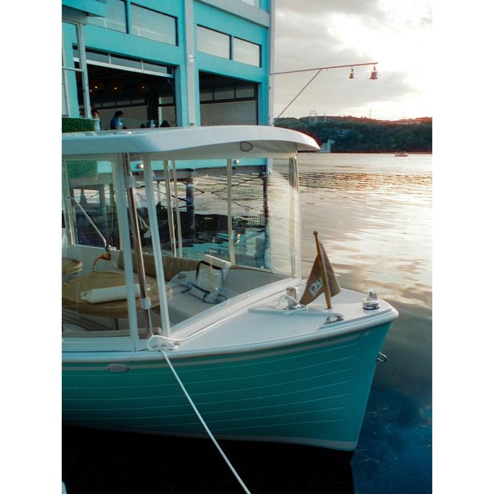 A turquoise boat is docked next to a building with a body of water and a distant shore visible under a cloudy sky.