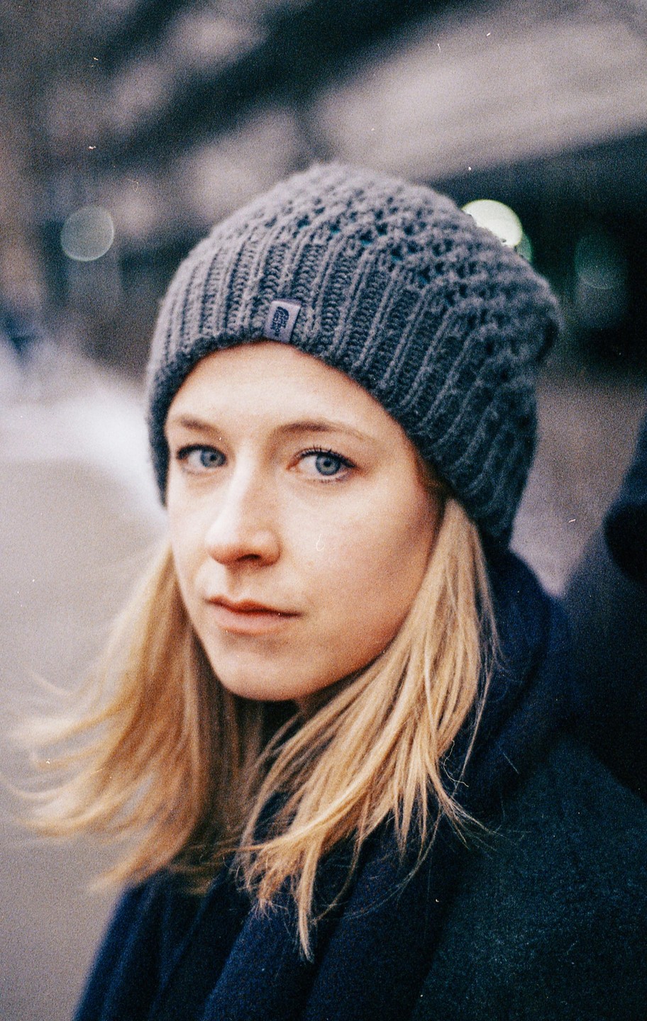 A woman with blonde hair and blue eyes wears a gray knit hat and a dark coat.