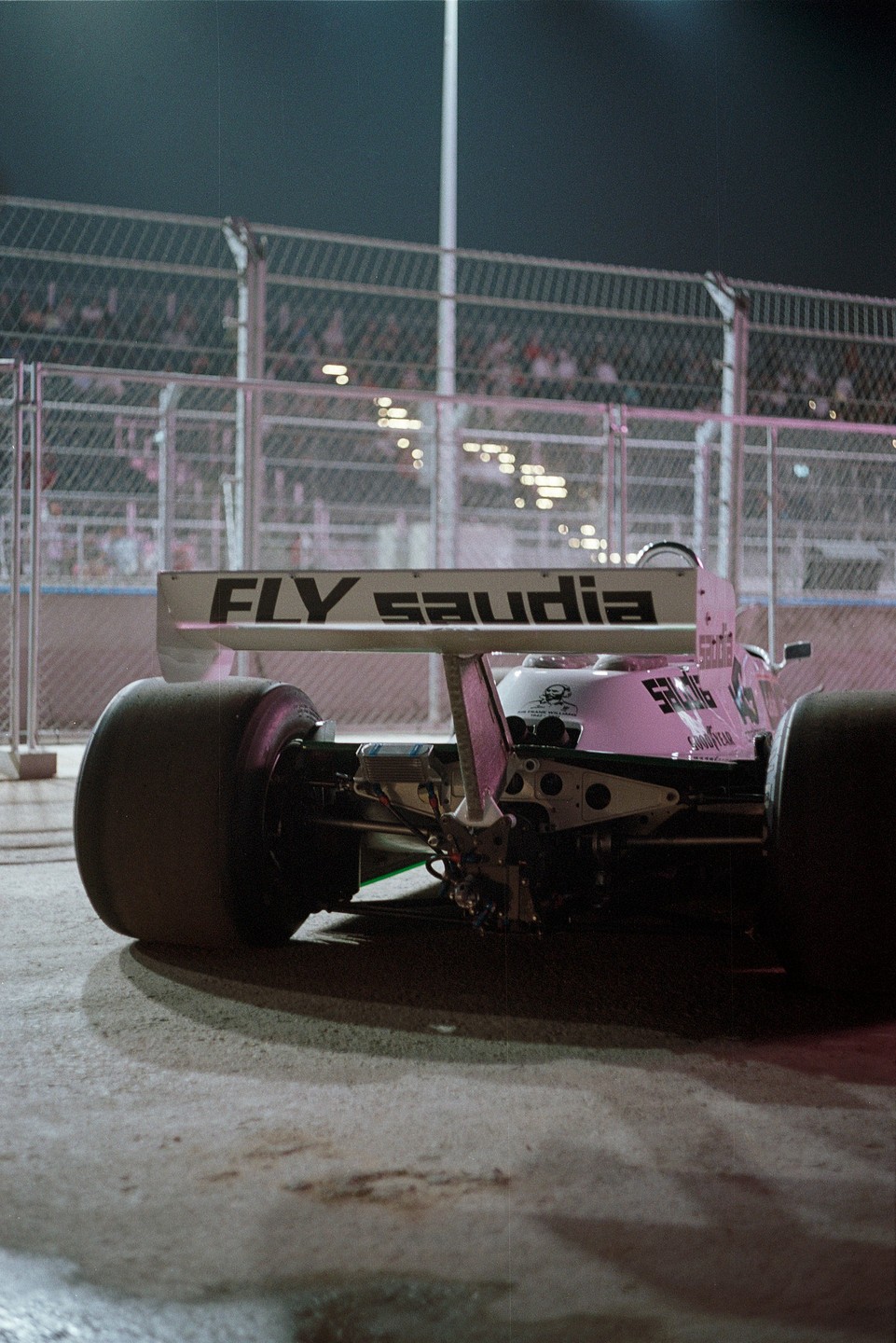 A racing car with "FLY saudia" written on its rear wing is parked on a track at night, with spectators visible behind a fence.
