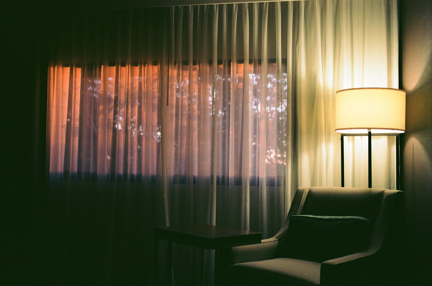 A window with sheer curtains shows an orange glow from outside, next to a floor lamp and an armchair.