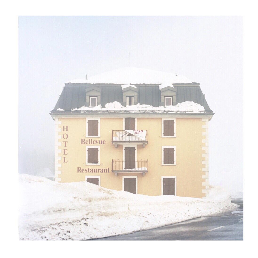 A snow-covered hotel with "Hotel Bellevue Restaurant" written on its facade stands beside a road with high snowdrifts.