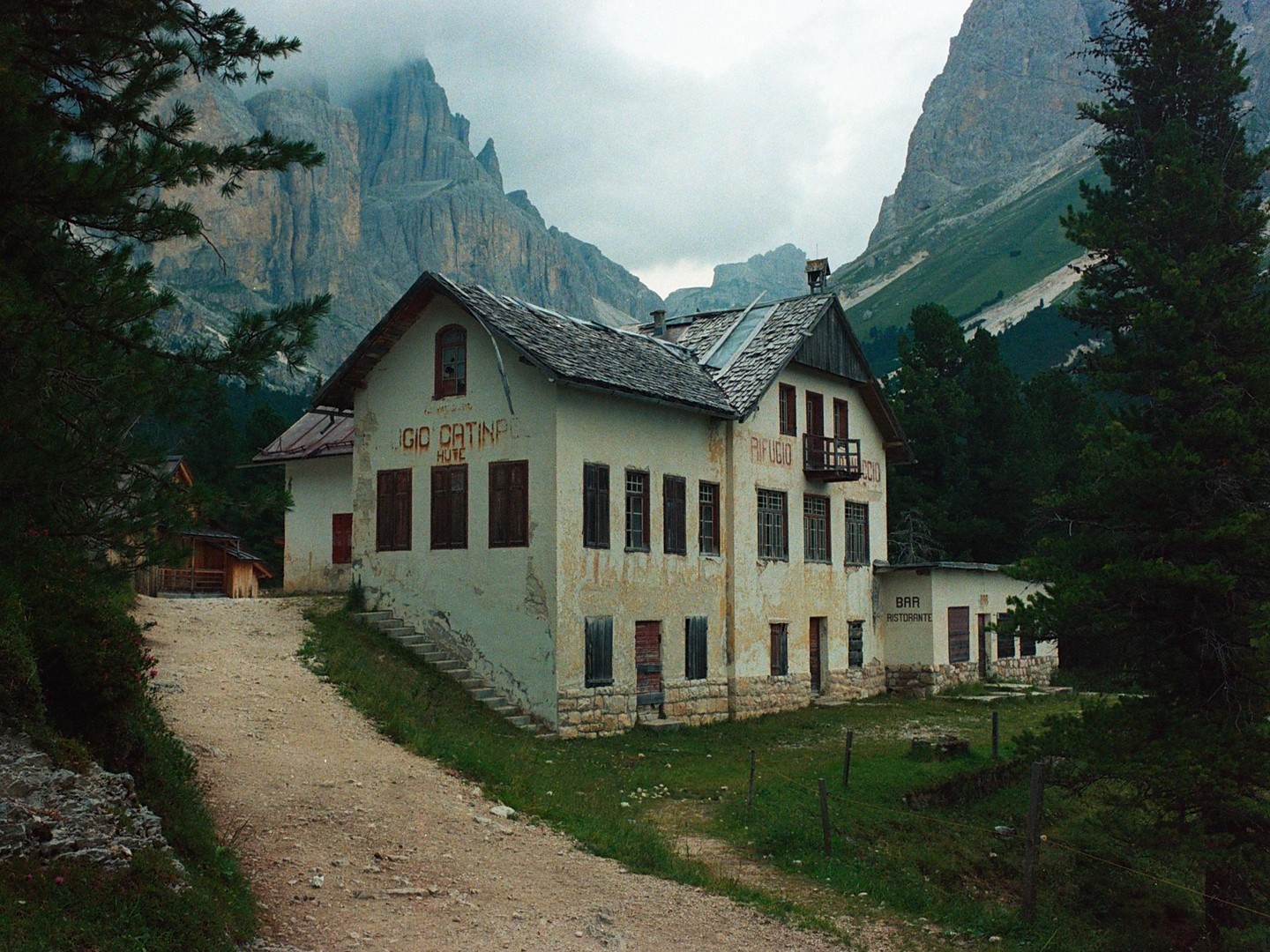 A dirt road leads to an abandoned hotel and restaurant building nestled in a valley surrounded by mountains and pine trees.
