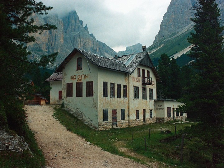 A dirt road leads to an abandoned hotel and restaurant building nestled in a valley surrounded by mountains and pine trees.