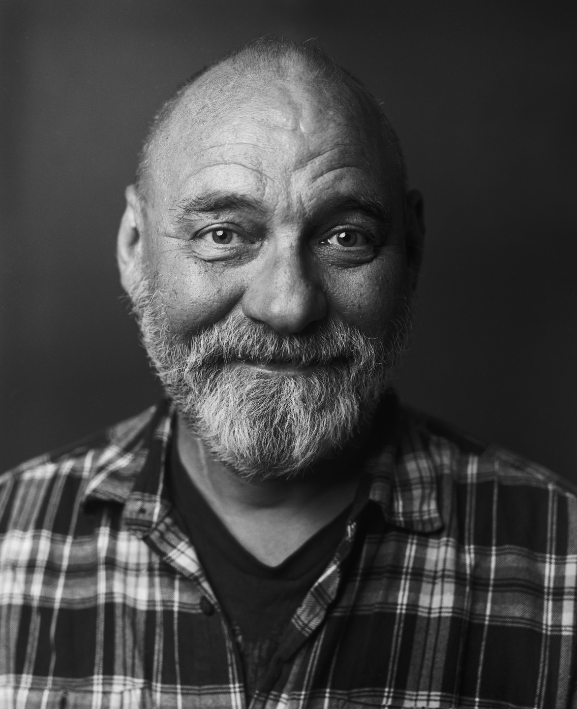 A man with a beard and a plaid shirt smiles at the camera in a black and white portrait.