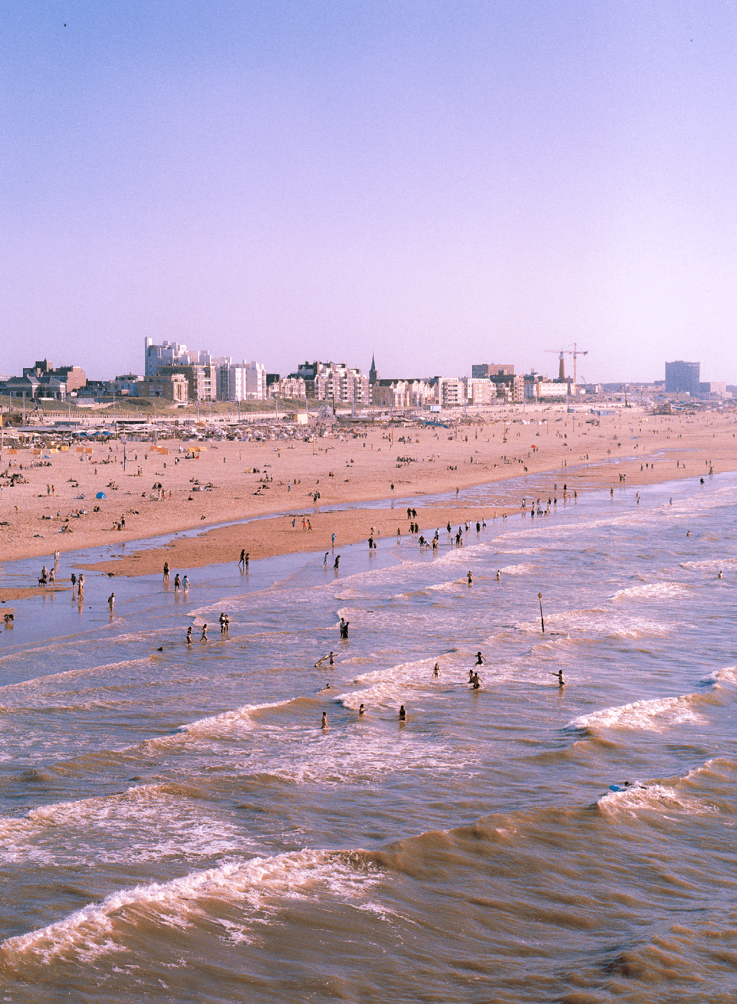 A wide shot shows a crowded beach with people in the water and on the sand, with a city skyline in the background.