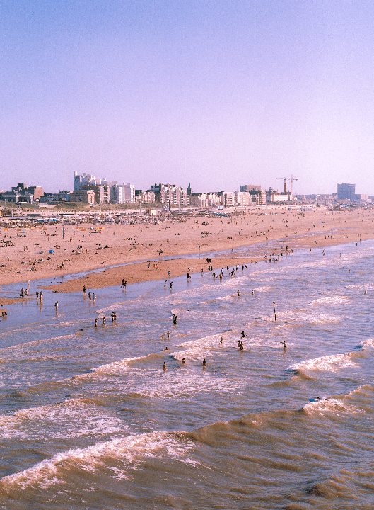A wide shot shows a crowded beach with people in the water and on the sand, with a city skyline in the background.