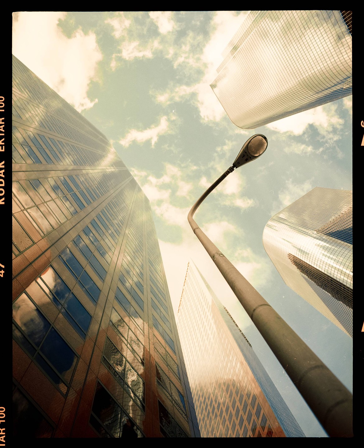 A low angle view shows a street light pole reaching towards the sky, surrounded by tall buildings with reflective windows.