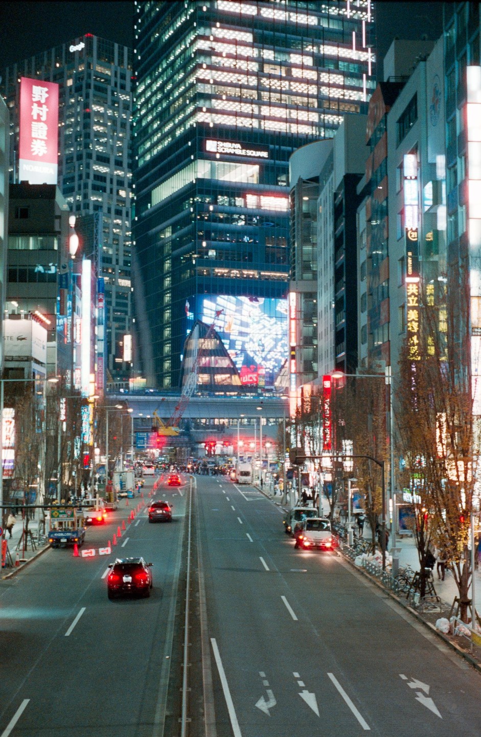 A wide street with cars and pedestrians is surrounded by tall buildings with illuminated signs and windows at night.