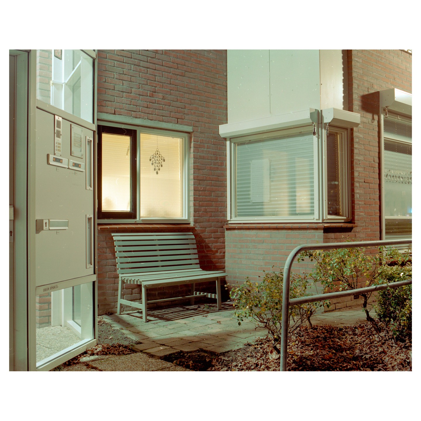 A light-colored bench sits on a brick patio next to a brick building with several windows, one of which is brightly lit from within.