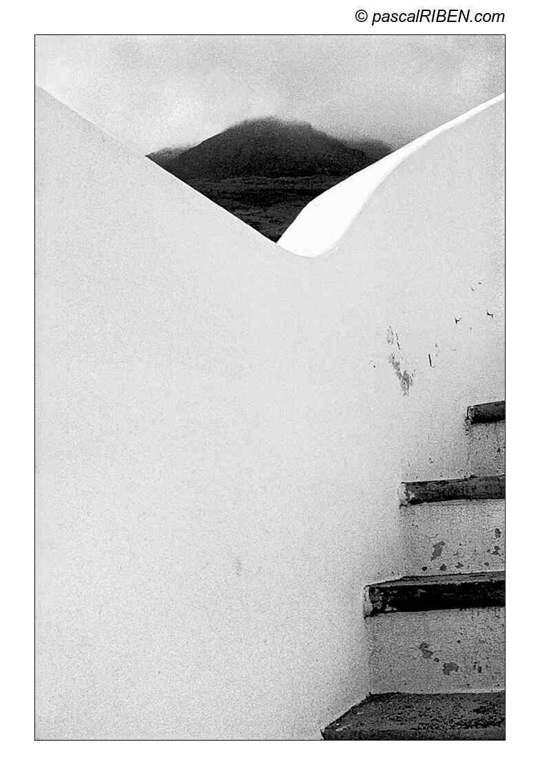 A black and white photo shows a mountain peak visible through a triangular opening in a white wall, with a staircase on the right.