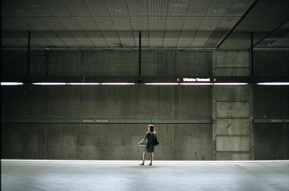 A woman stands alone on a subway platform with "Wilshire/Vermont" signs visible.