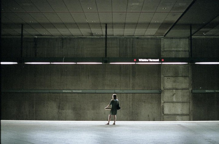 A woman stands alone on a subway platform with "Wilshire/Vermont" signs visible.