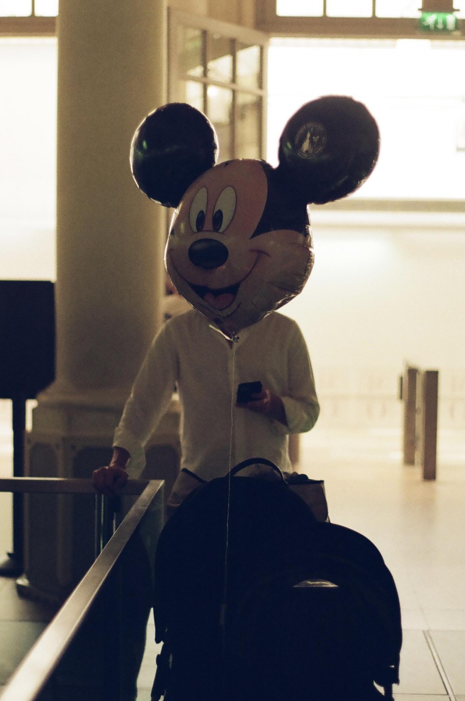 A person with a Mickey Mouse balloon head stands in a bright room with a backpack in the foreground.