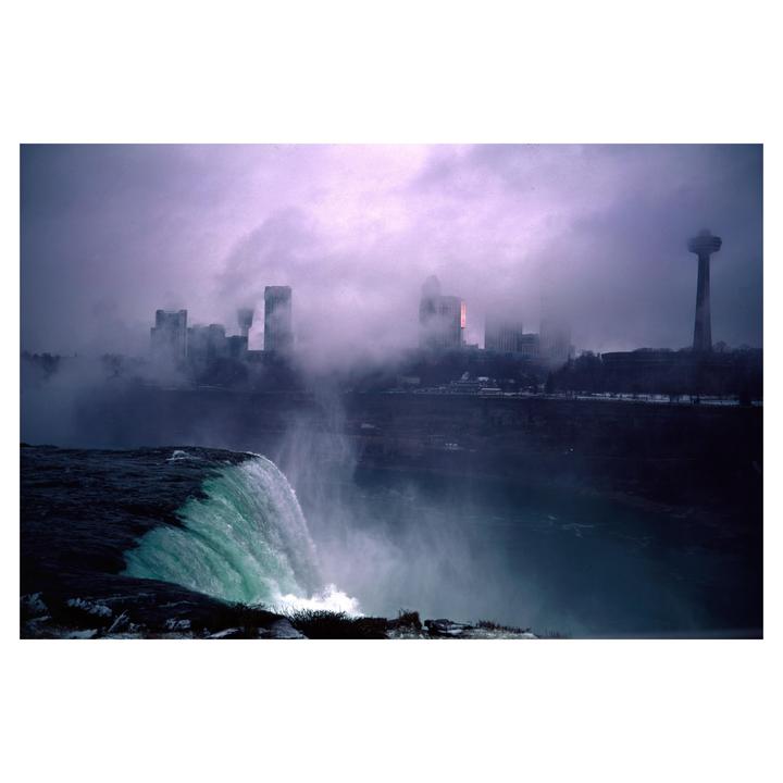 A waterfall cascades into a river with a city skyline and tower visible through heavy fog in the background.
