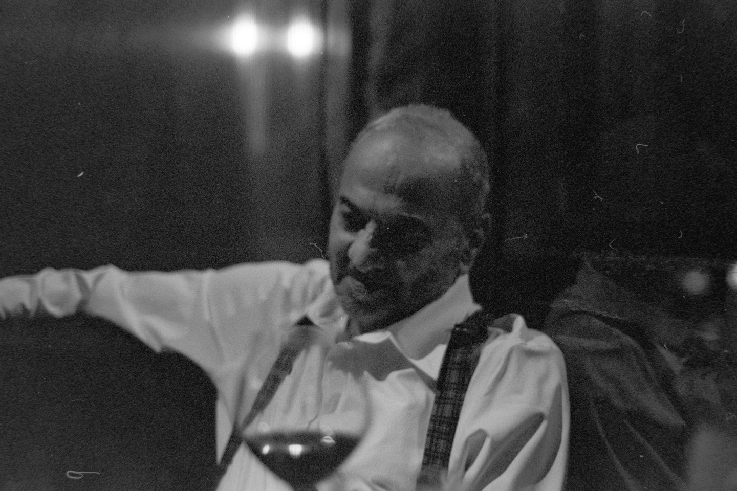 A man in a white shirt and suspenders sits with a glass of wine in a dimly lit room.