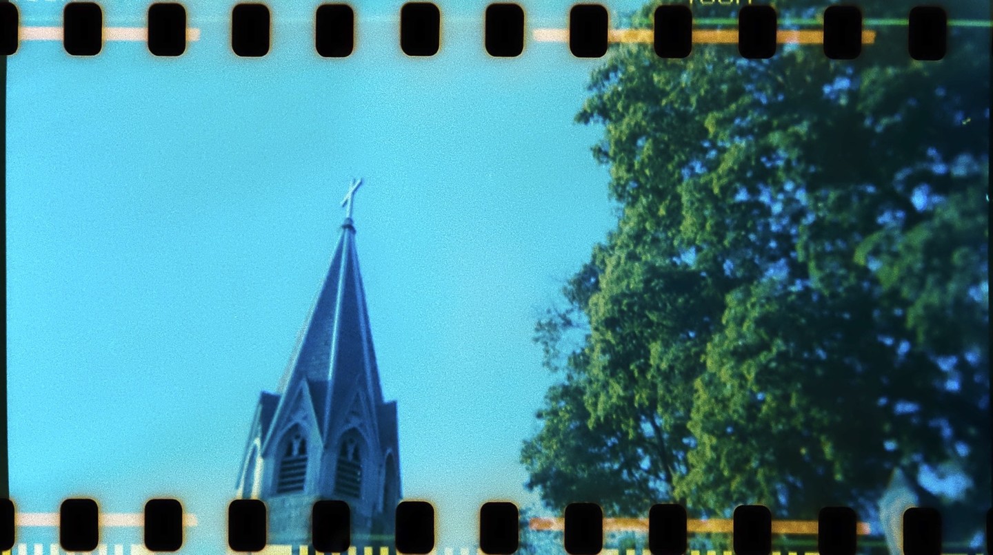 A church steeple with a cross on top stands against a clear blue sky next to a large green tree.