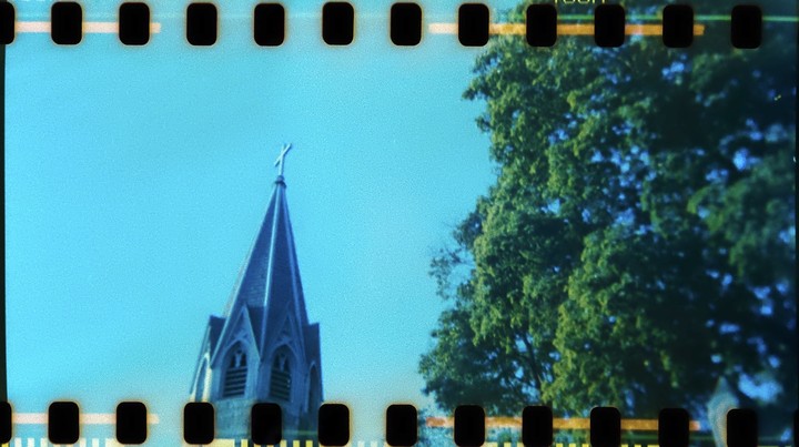 A church steeple with a cross on top stands against a clear blue sky next to a large green tree.