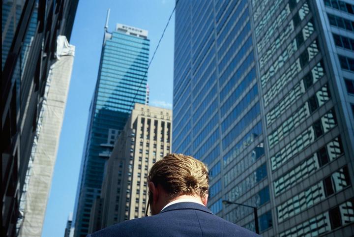 A man in a suit stands looking up at tall buildings in a city.