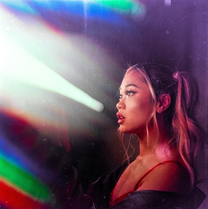 A woman with pigtails looks to the left with a light beam and rainbow flare in the foreground.