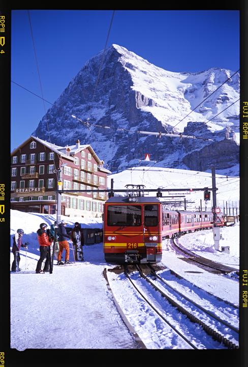 A red train is stopped on a snowy track next to a building with a large snow-covered mountain in the background.