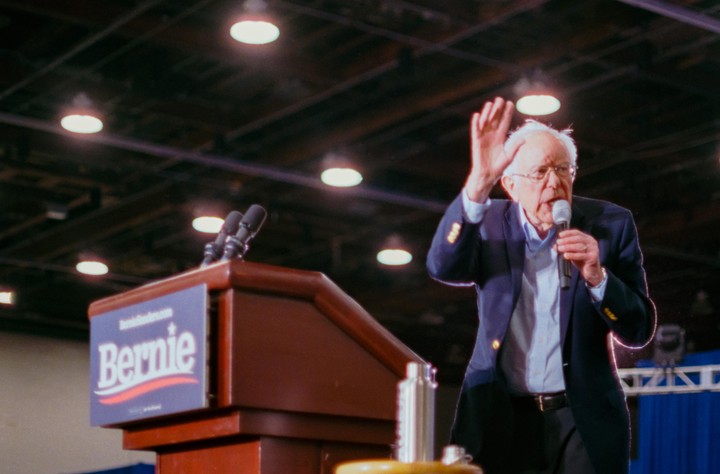 A man in a suit and glasses speaks into a microphone with one hand raised, standing next to a podium with a "Bernie" sign.