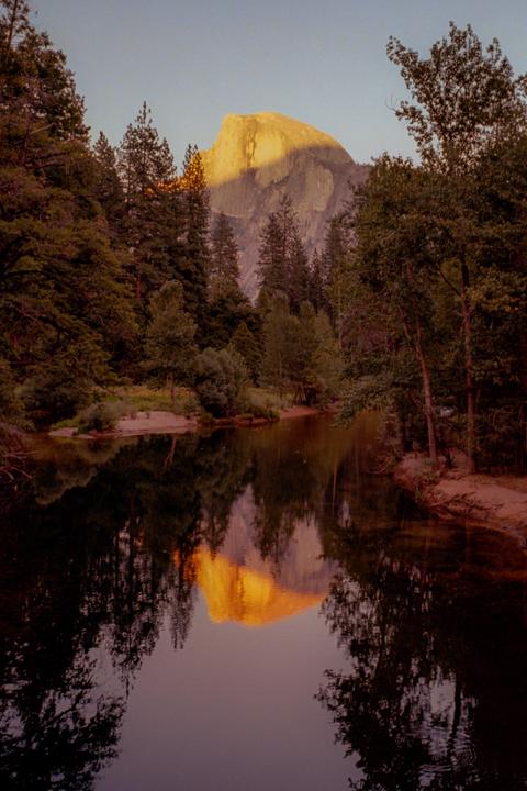 A mountain with a sunlit peak is reflected in a body of water surrounded by trees.
