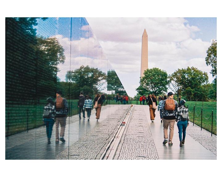 People walk along a path next to a reflective wall with the Washington Monument in the distance.