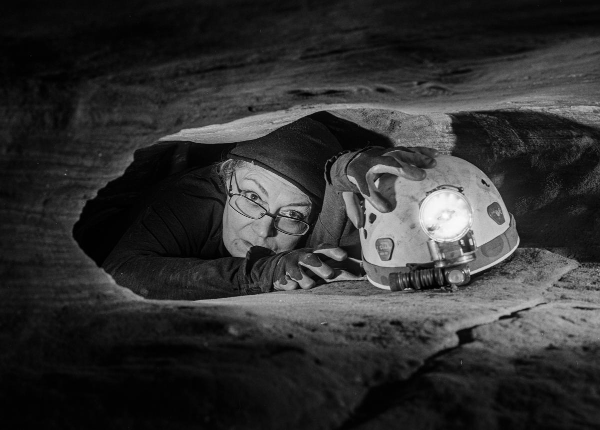A woman wearing glasses and a hat is crawling through a narrow cave opening, holding a helmet with a bright headlamp.