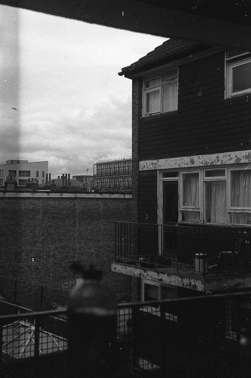 A black and white image shows a building with a balcony and a cityscape in the background under a cloudy sky.