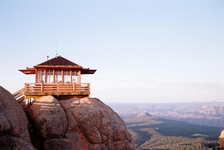 A fire lookout tower sits atop a rocky peak overlooking a vast forest and mountain range.