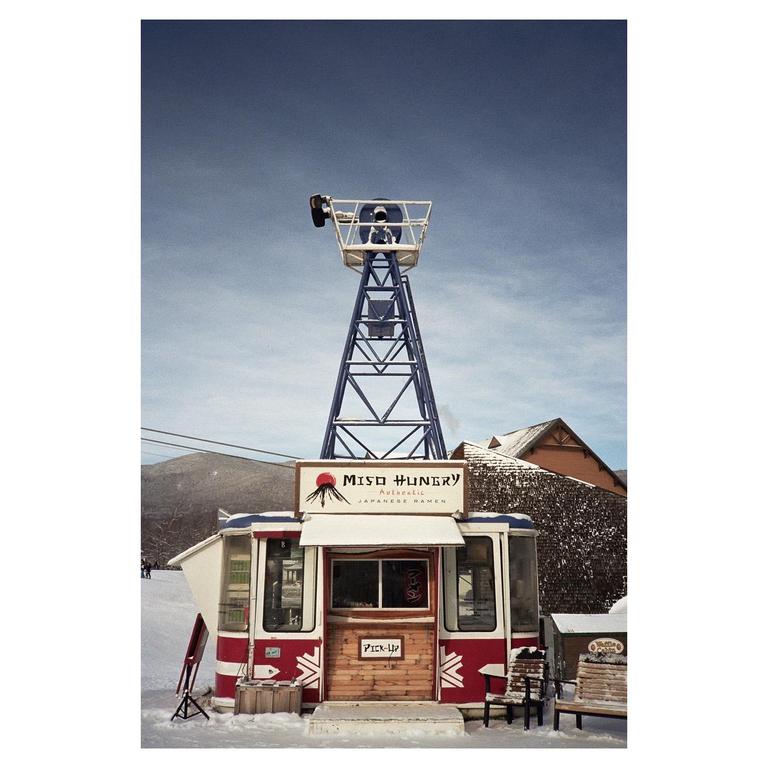 A red and white ramen food truck sits in the snow beneath a tall blue tower.