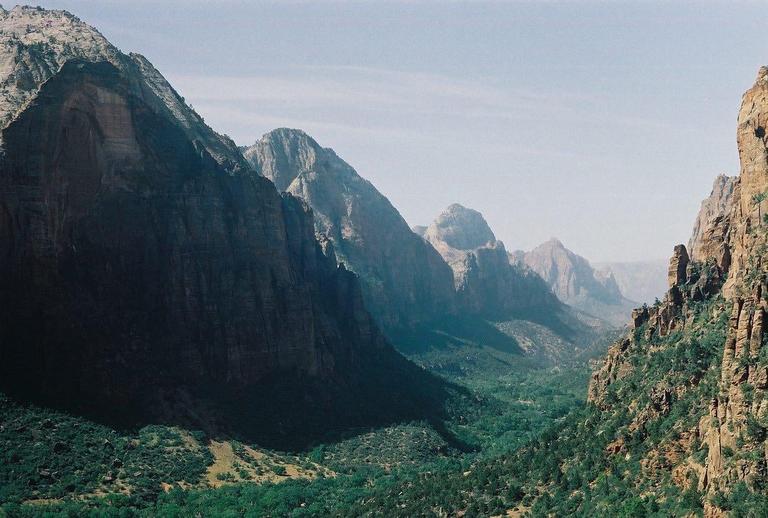 A valley with green trees and bushes is surrounded by tall, rocky mountains under a hazy sky.