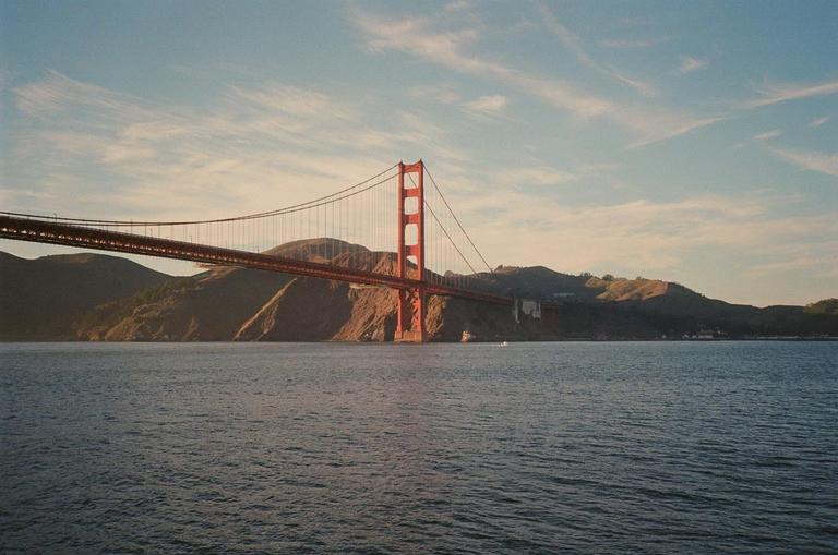 The Golden Gate Bridge spans across a body of water with hills in the background under a partly cloudy sky.