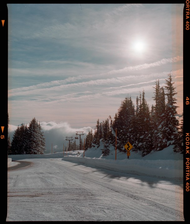 A snow-covered road curves through a forest of pine trees under a bright sun and cloudy sky.