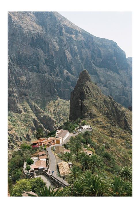 A small village with a winding road is nestled in a valley surrounded by steep, rocky mountains and palm trees.