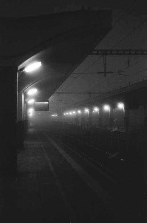 A train station platform is illuminated by overhead lights on a foggy night.
