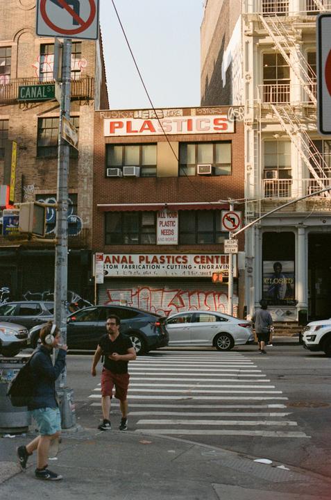 A man and a woman cross a street at an intersection with cars and buildings in the background.